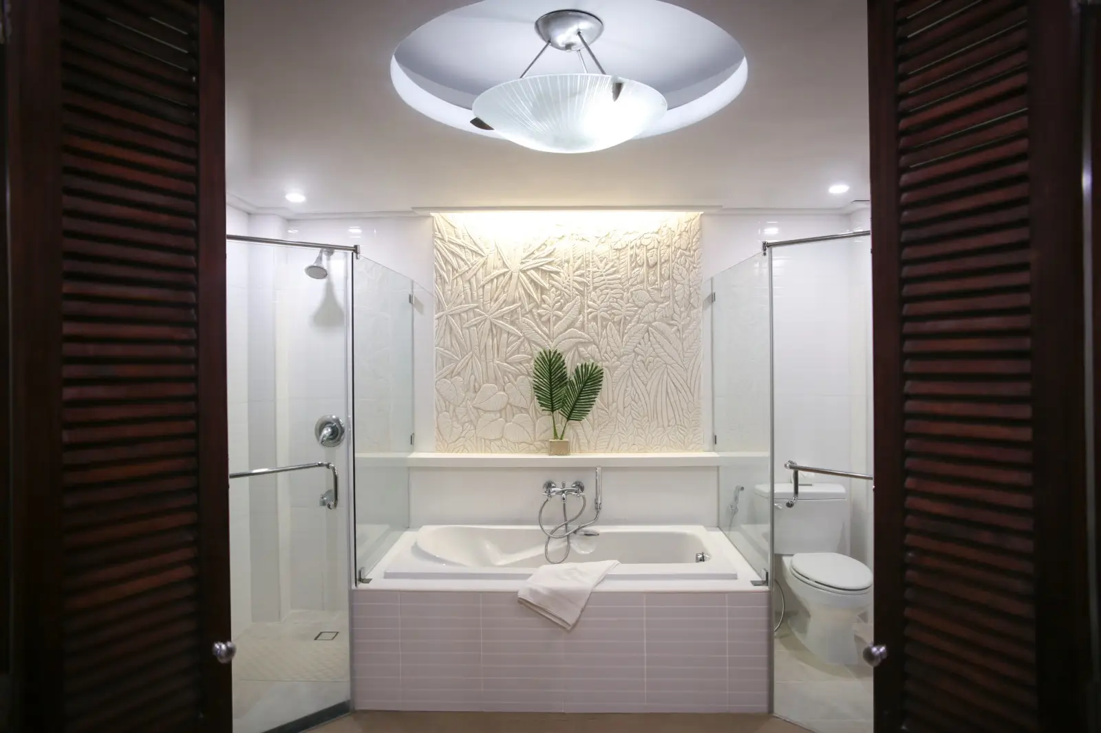 Spacious modern bathroom in Mandarin Bay Resort & Spa featuring a large white bathtub with an intricately carved tropical design on the wall above, and a separate glass shower enclosure.