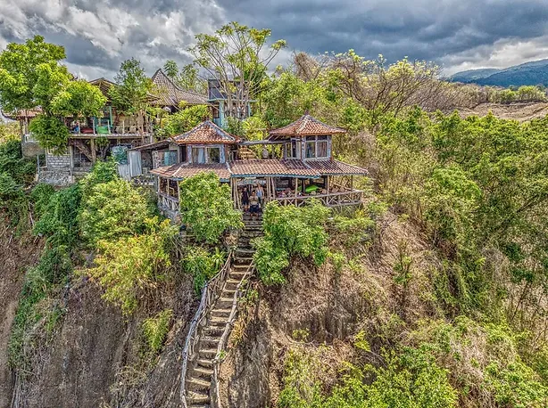 Aerial view of Santhika Retreat Center, perched on a lush cliffside in Bali, featuring multiple traditional Balinese pavilions connected by rustic wooden stairways. 