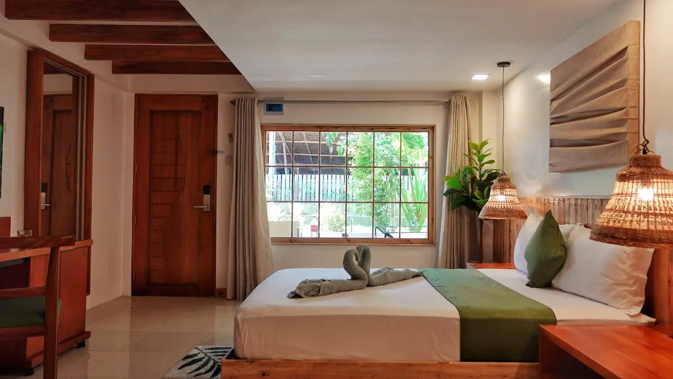 Inviting guest room at Senare Boracay Hotel, decorated with a large bed featuring a creatively folded towel art, wooden furniture, woven lampshades, and a view of the outdoors through a large window, creating a cozy and welcoming atmosphere.