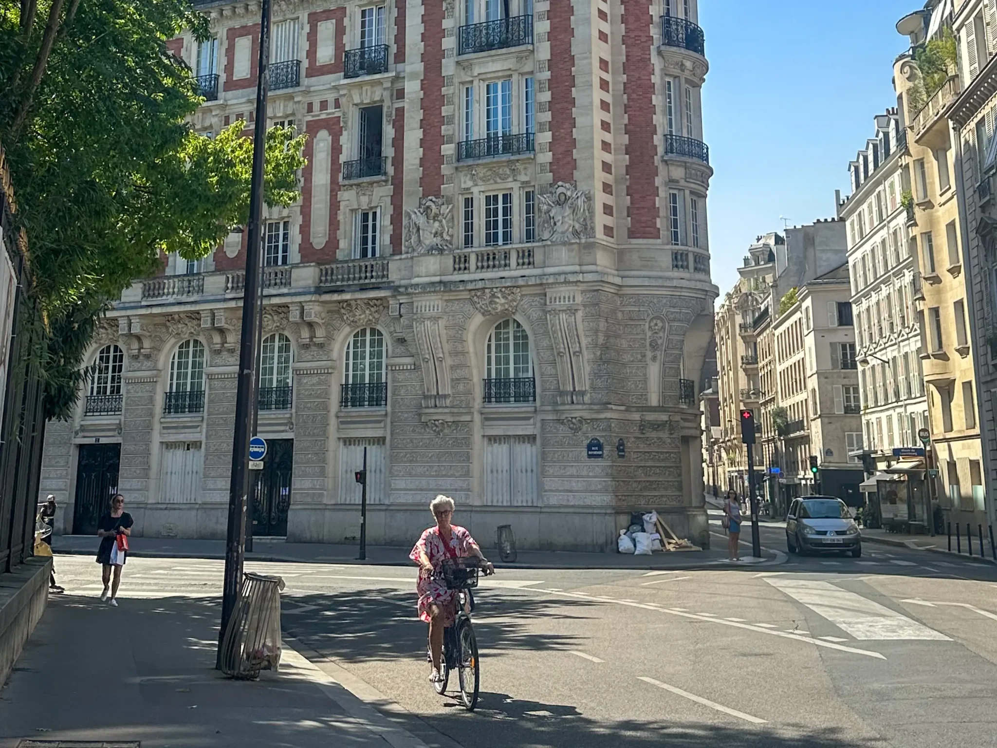 A slow travel in Paris featuring an elderly woman cycling past a historic building with striking red and white facade. The street is quiet with a few pedestrians and parked cars, framed by lush green trees and clear blue skies.