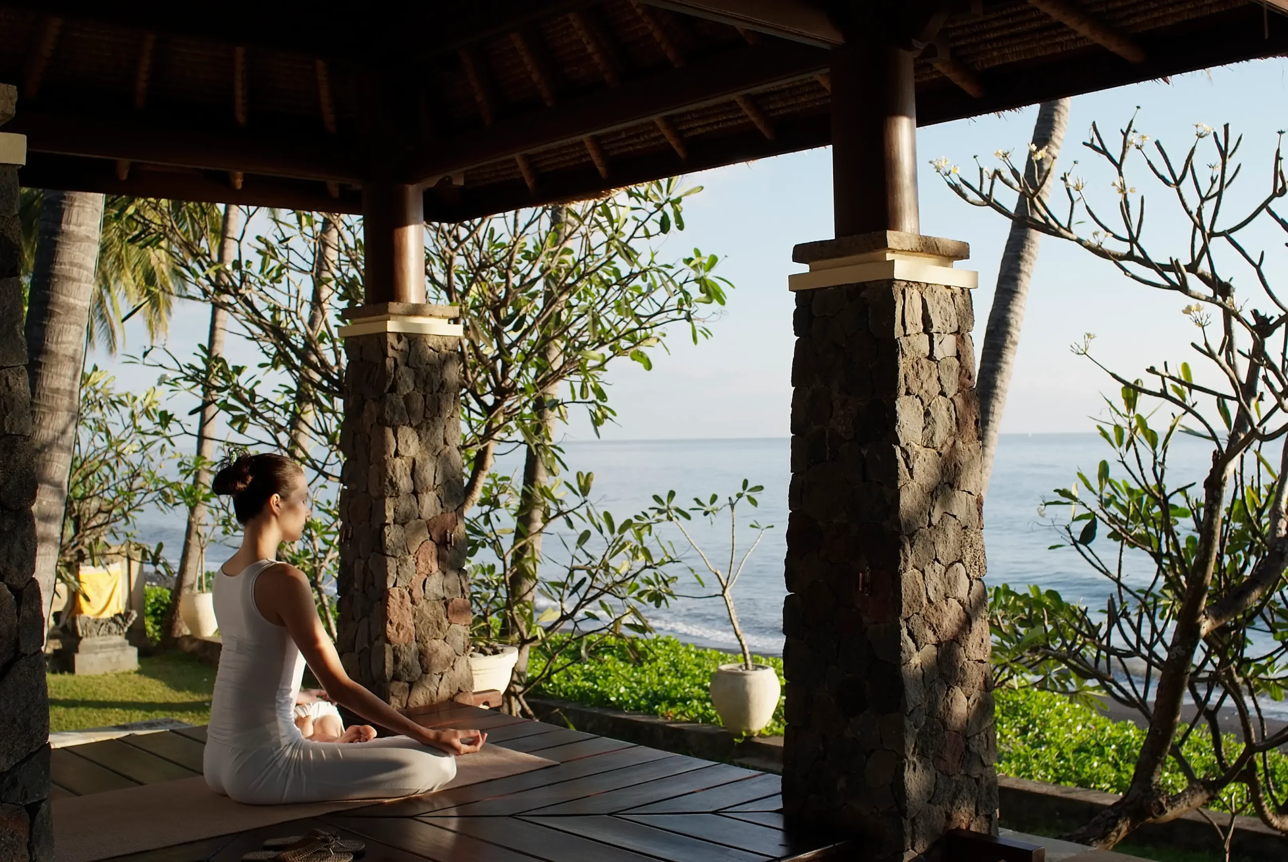 A woman in white yoga attire practices meditation at a wellness resort in Bali, within a seaside yoga pavilion.