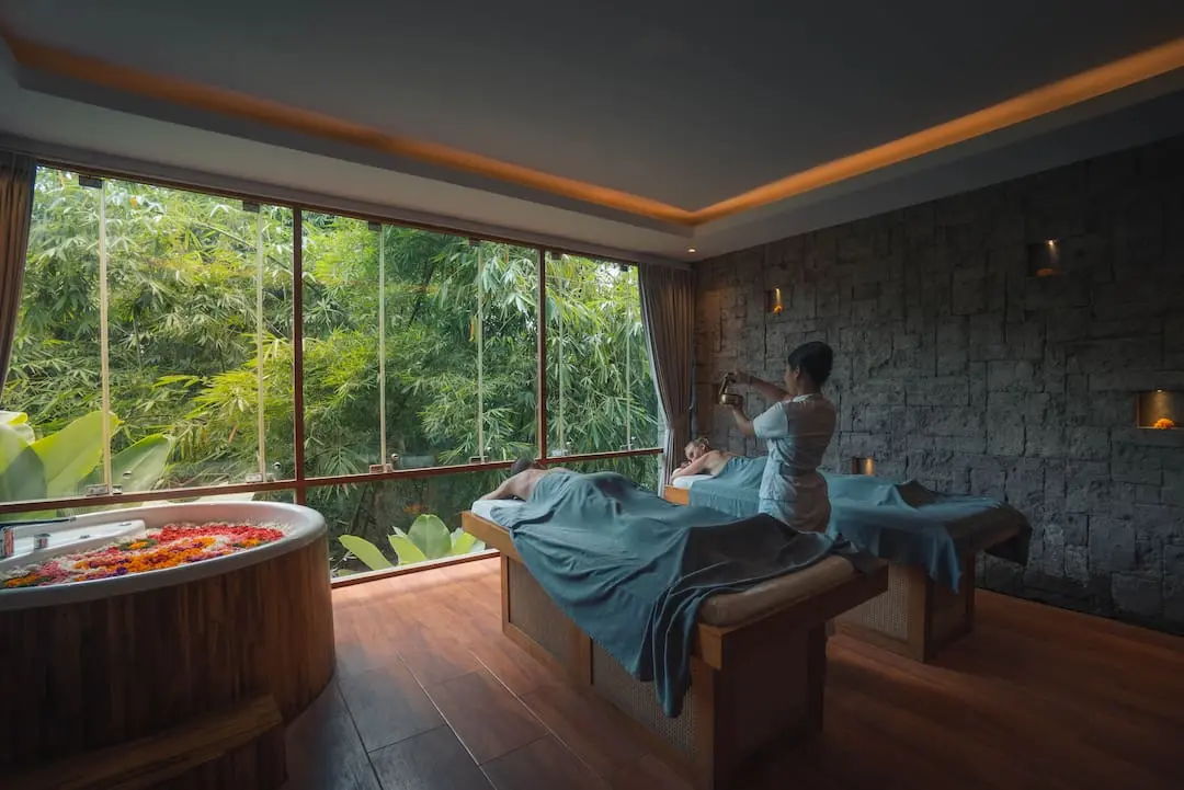 Interior of a luxury spa room featuring two massage tables with blue sheets and a therapist in action. The room has a panoramic window showcasing dense tropical foliage outside, providing a tranquil atmosphere.