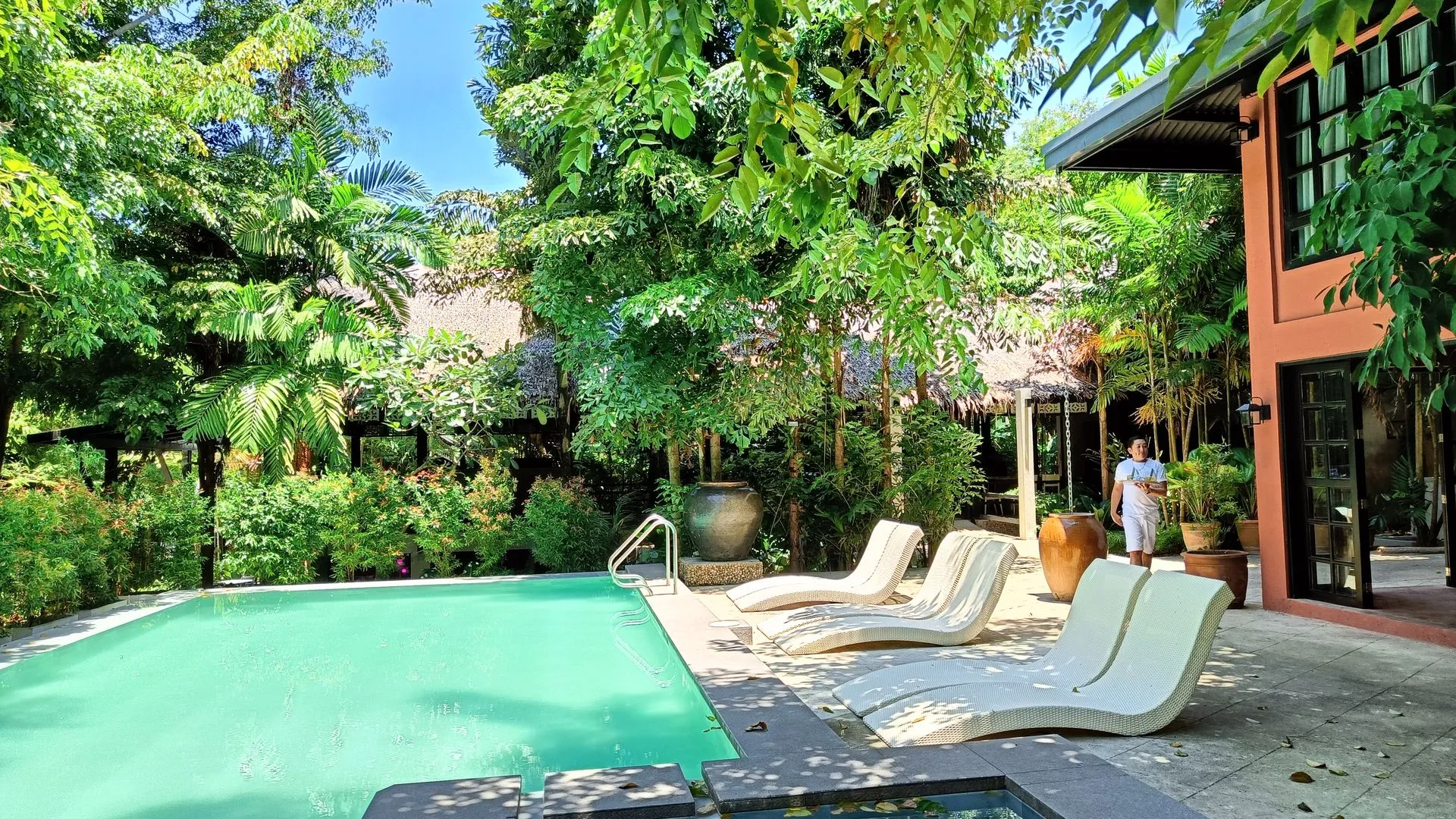 Lush poolside view at The Henry Resort Boracay, a boutique hotel in Boracay, featuring vibrant greenery, modern sun loungers, and a tranquil swimming pool adjacent to a rustic style building.