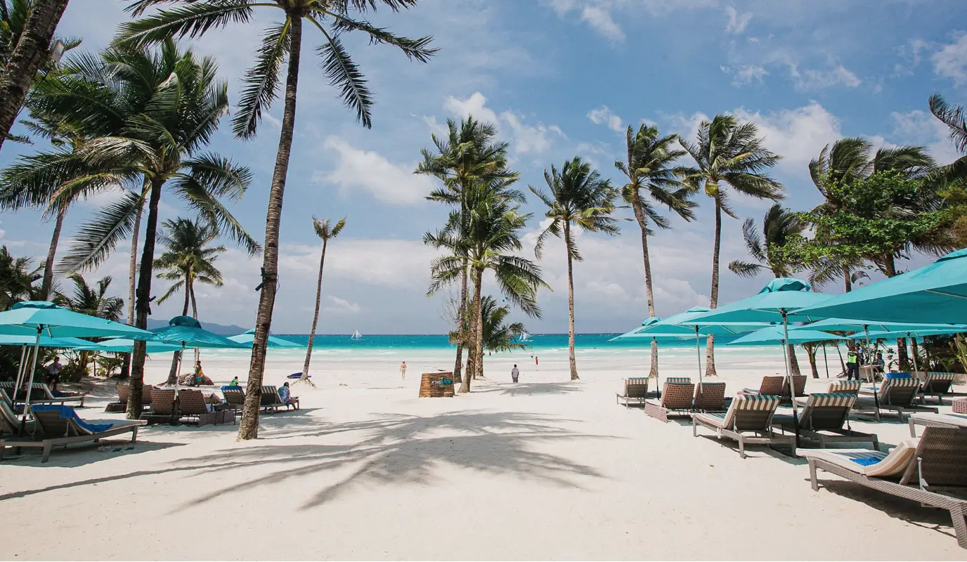 Scenic view of the pristine beach at The Lind Boracay, a Boracay boutique resort, lined with tall palm trees and turquoise beach umbrellas, overlooking the crystal-clear waters.
