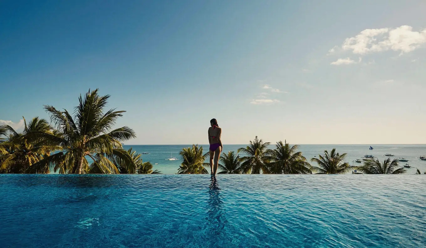 A person stands contemplatively at the edge of an infinity pool at  The Lind Boracay, a boutique hotel in Boracay, enjoying the panoramic view of palm trees and the sea under a clear sky.