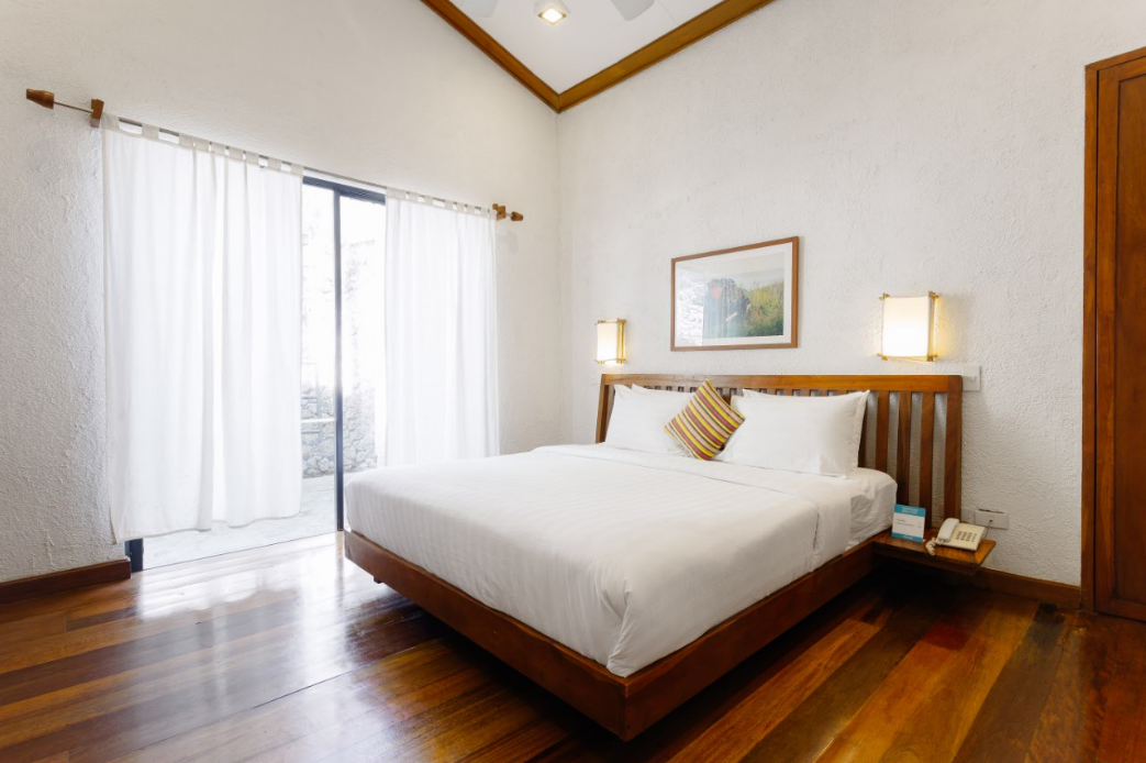 Elegantly simple bedroom at The Strand Boutique Resort, Boracay, with a king-size bed, polished wooden flooring, white walls, and a framed picture above the bed, bathed in natural light from large glass doors leading to an outdoor area.