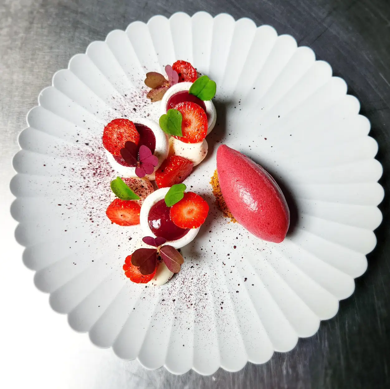 A beautifully plated dessert at Tomy & Co with strawberries, meringue, and a scoop of sorbet on a white fluted plate.