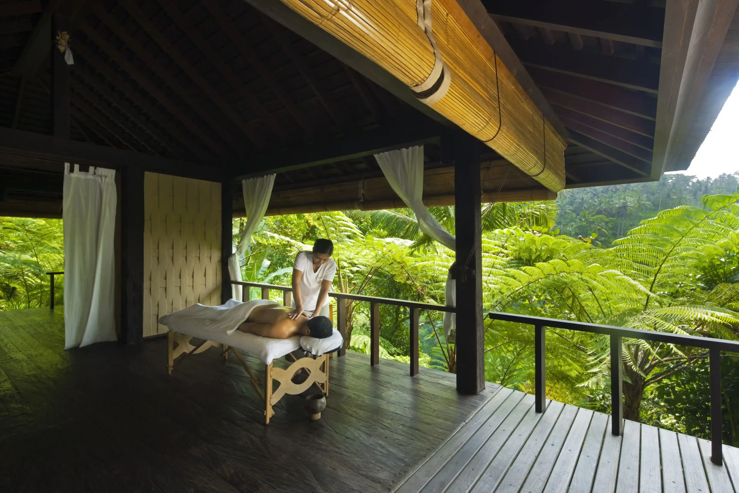 A therapeutic massage session at a wellness resort in Bali, conducted on a covered balcony set against a backdrop of lush tropical vegetation.