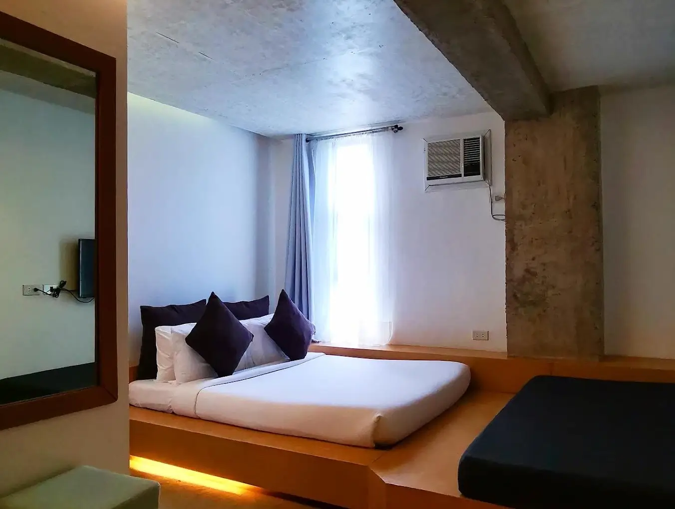pacious room at Urban Boutique Hotel, Boracay, with a queen-sized bed set on a lighted platform, concrete walls, a large mirror, and a cushioned bench, illuminated by natural light from the window.