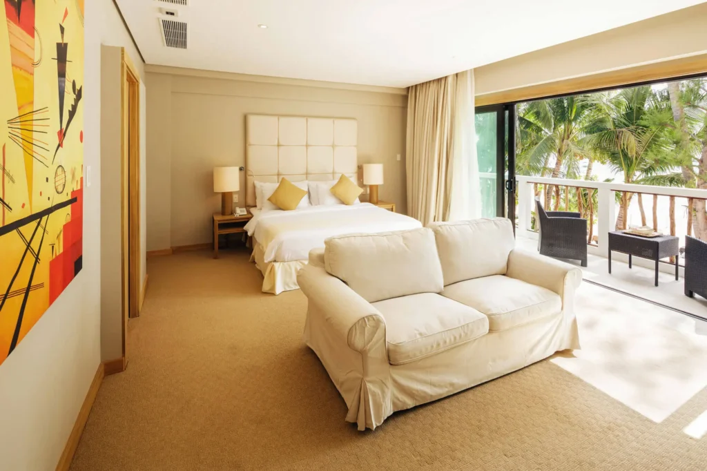 Luxurious and spacious bedroom at Villa Caemilla Beach Boutique Hotel in Boracay. The room features a large bed with a cushioned headboard, an elegant sofa, and opens onto a balcony with a view of palm trees.