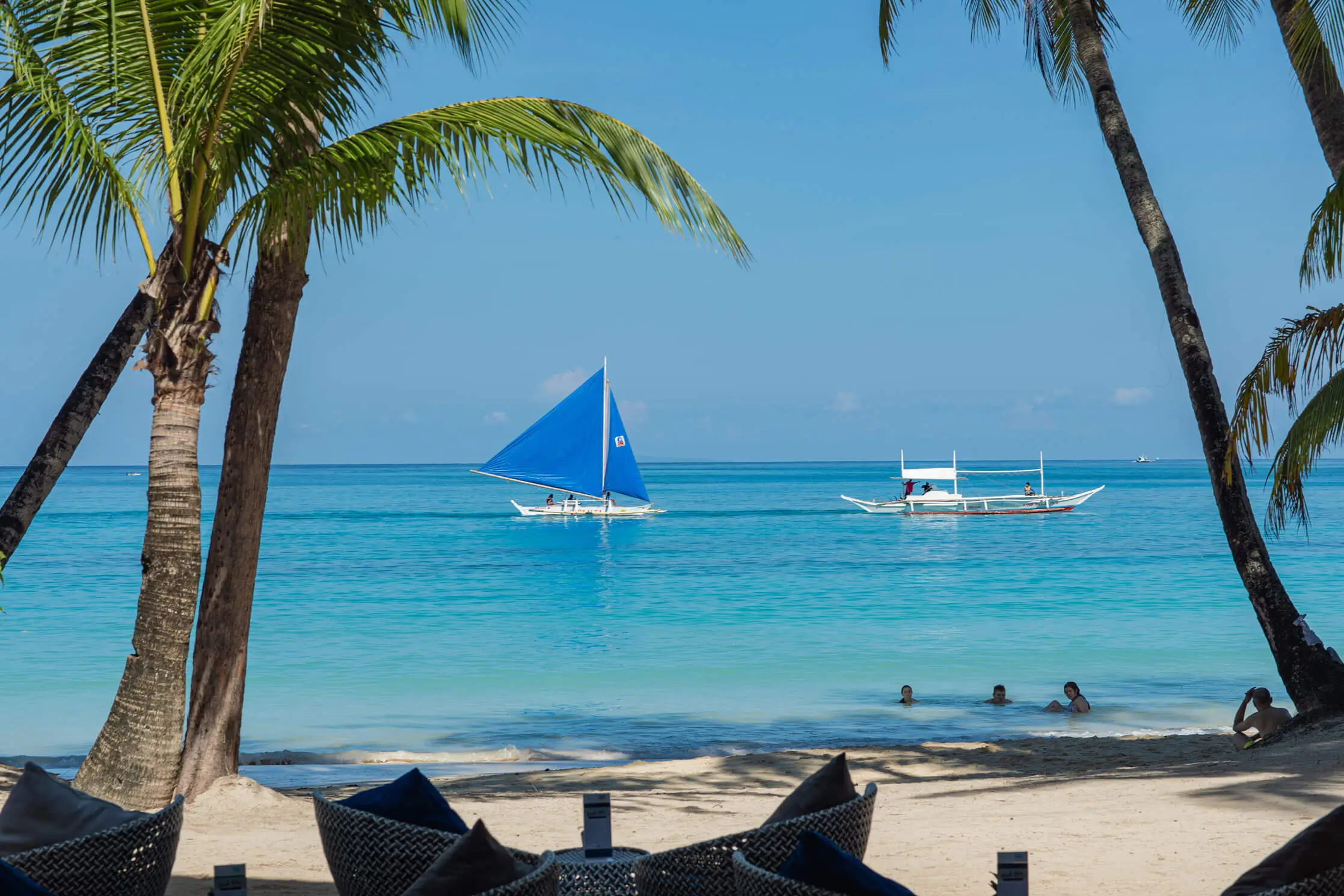 Idyllic beachfront view at Villa Caemilla Beach Boutique Hotel in Boracay, showcasing a traditional sailboat on the clear blue waters. The beach is lined with palm trees and inviting lounge chairs, perfect for relaxation.