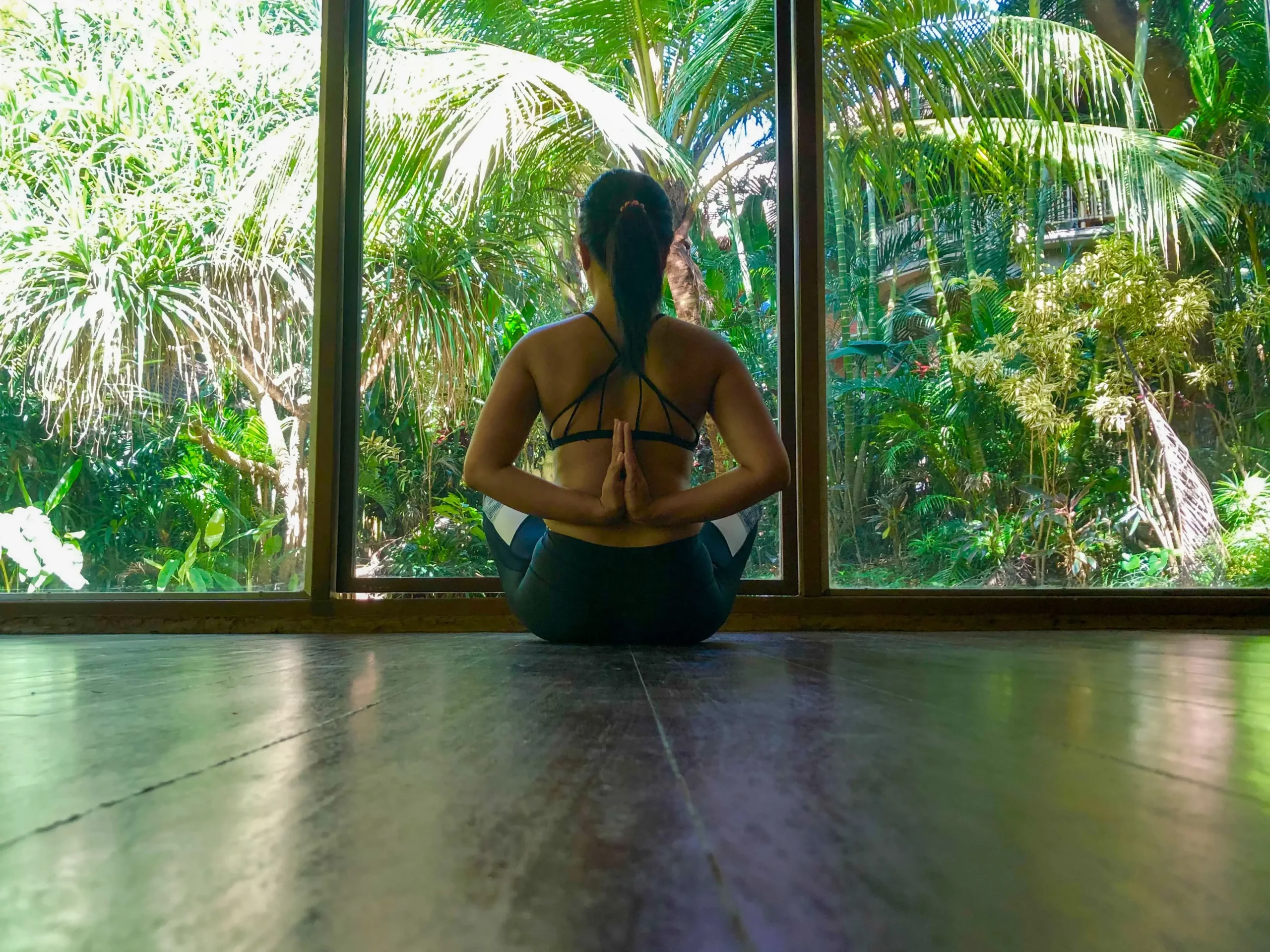  practice yoga in a seated twist pose, facing a large window overlooking a lush tropical garden at Yoga Barn Retreat in Bali. 