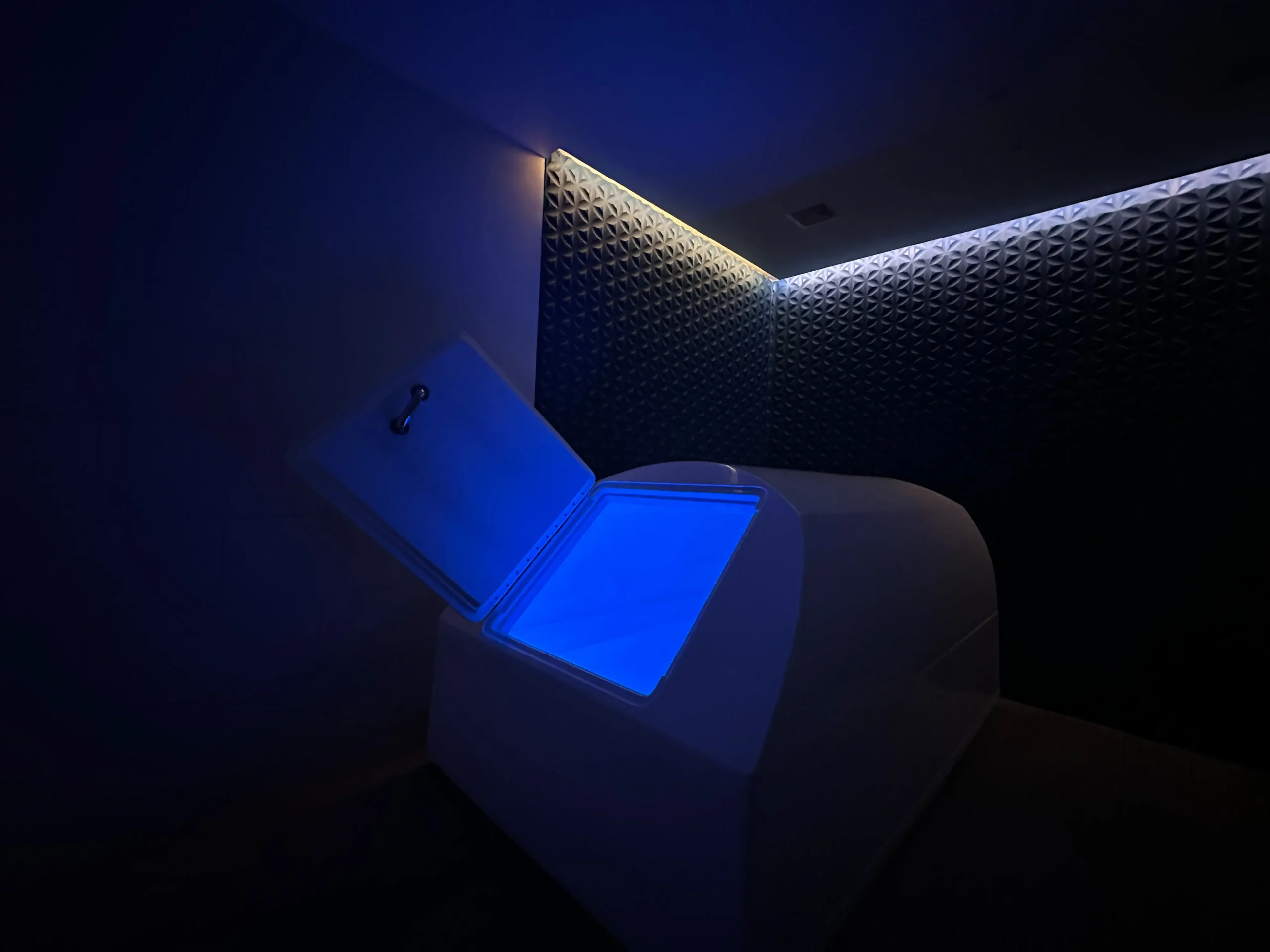 The exterior view of a modern zero-gravity flotation therapy tank in a dimly lit room with textured walls, set in Solace Float. This tank is designed for deep relaxation, reducing sensory input to a minimum to help achieve a meditative state.