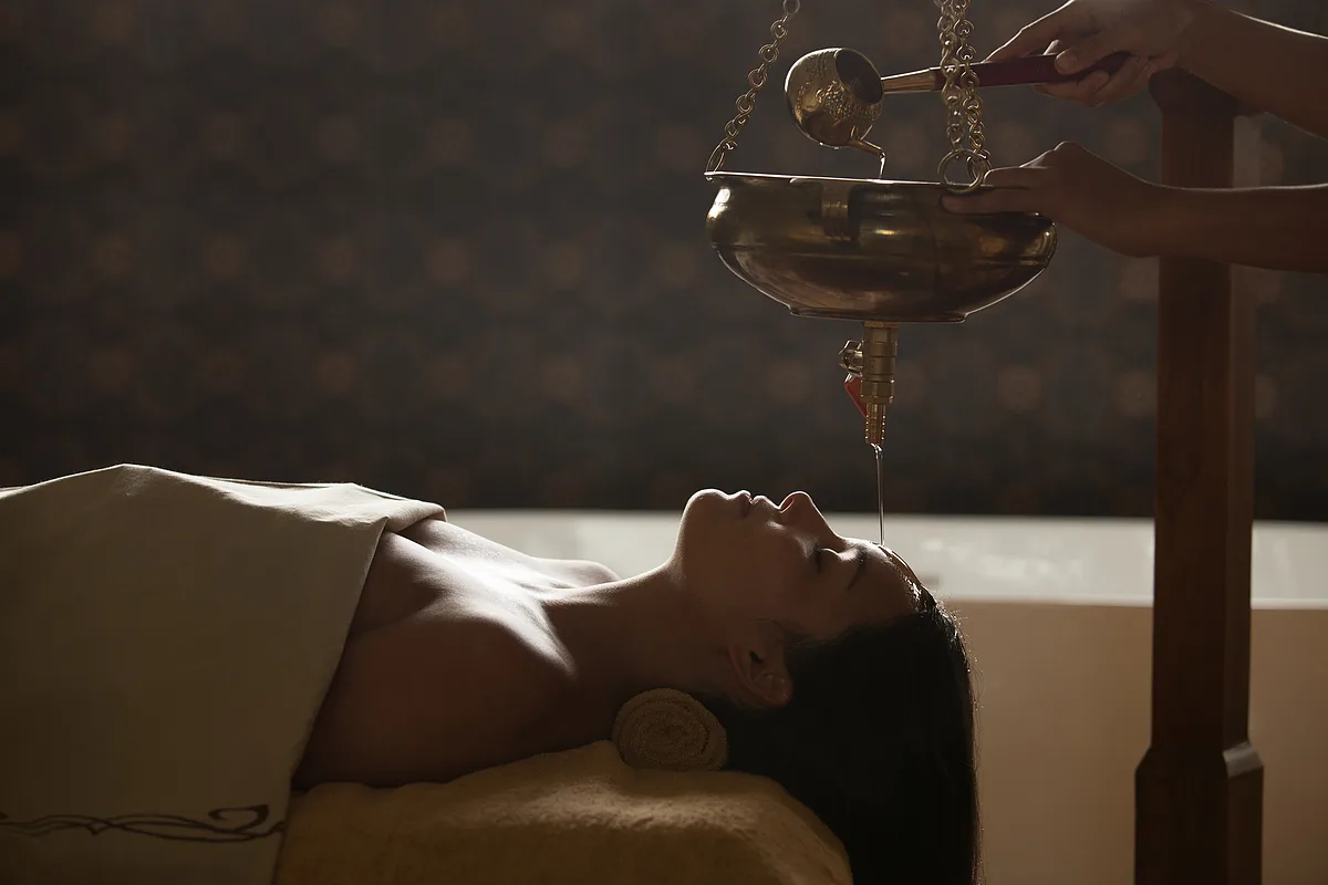 Woman lying down receiving a Shirodhara treatment, with warm oil poured onto her forehead from a hanging brass bowl, at a luxury wellness retreat in Ubud, Bali