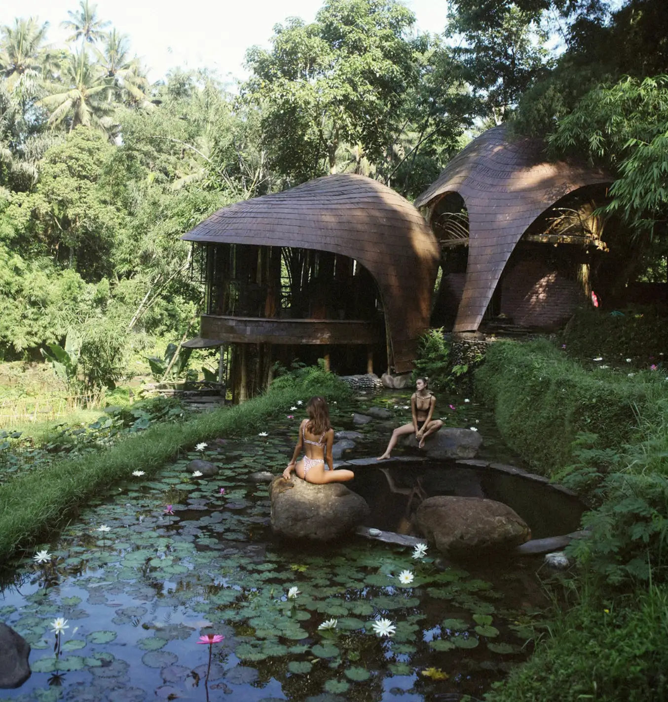 Two women in bikinis sit on large rocks by a lily pond, surrounded by lush greenery. In the background, there is Bambu Indah Bali, a unique bamboo structure with curved, overlapping roofs