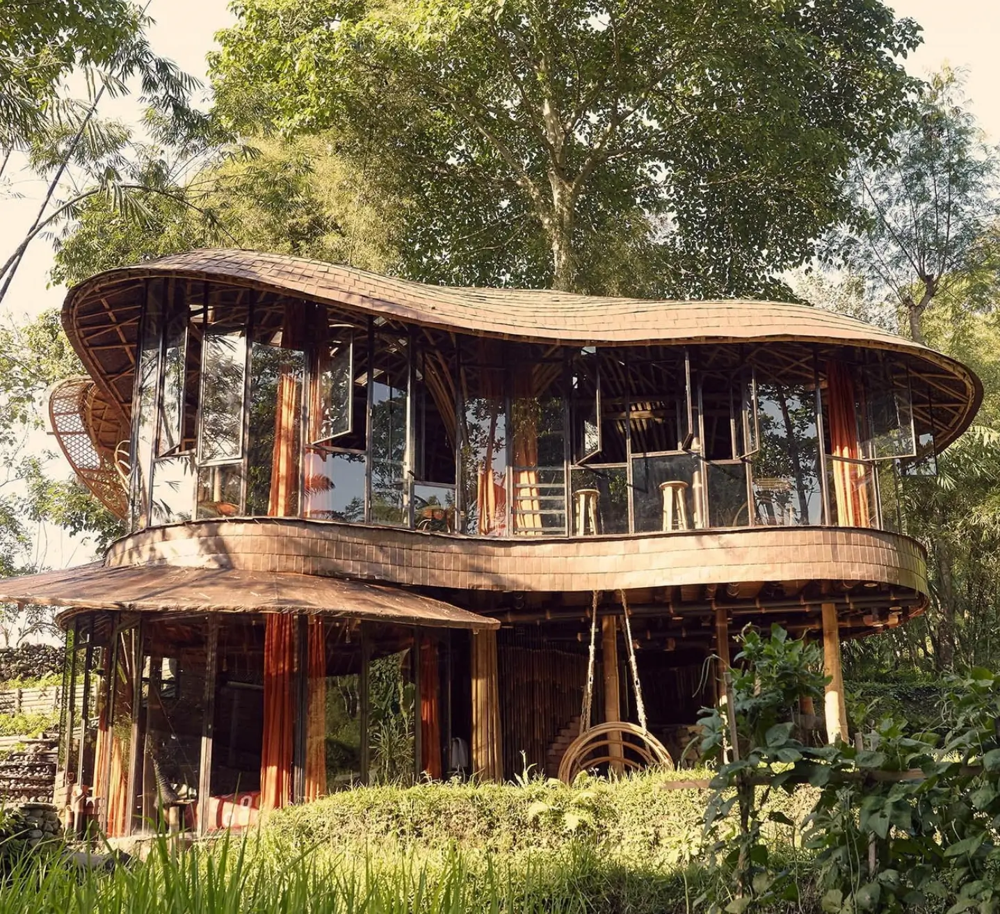 Bambu Indah, beautifully designed bamboo house with large, curved roofs and extensive windows, set amidst lush green trees.