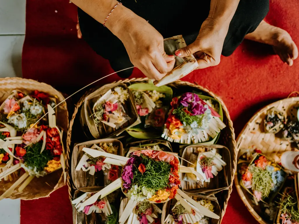 Hands preparing canang sari, Balinese offerings of flowers and incense in a small basket made of palm leaves.