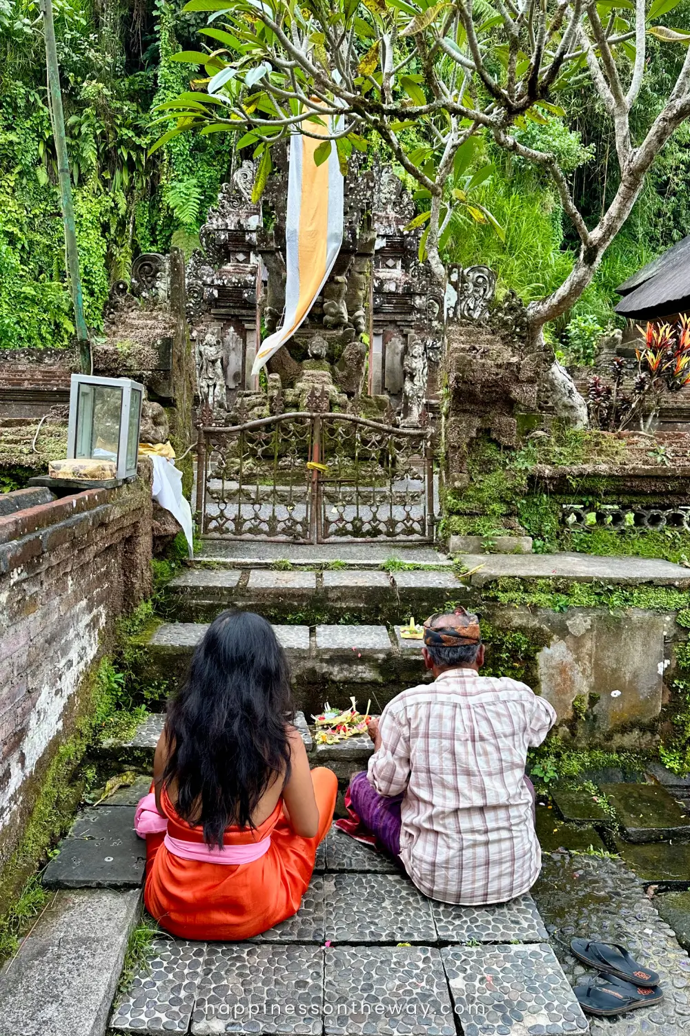 I am wearing an orange sarong with a Balinese priest sitting and praying at an ancient, moss-covered Balinese temple, Pura Gunung Kawi Sebatu, one of the hidden gems in Ubud.