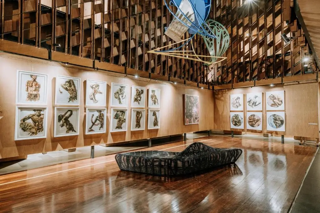 An art gallery at Artotel Sanur with wooden walls featuring multiple framed paintings depicting abstract human figures. A black, woven lounge chair sits in the center of the polished wooden floor, and a modern sculpture hangs from the ceiling above.