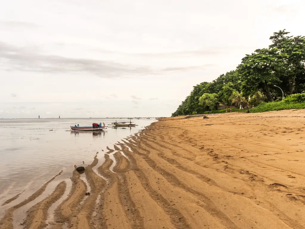 A tranquil view of Karang Beach in Sanur, Bali, with shallow water creating rippled patterns in the sand, a few small boats floating near the shore, and lush green trees lining the edge of the beach.