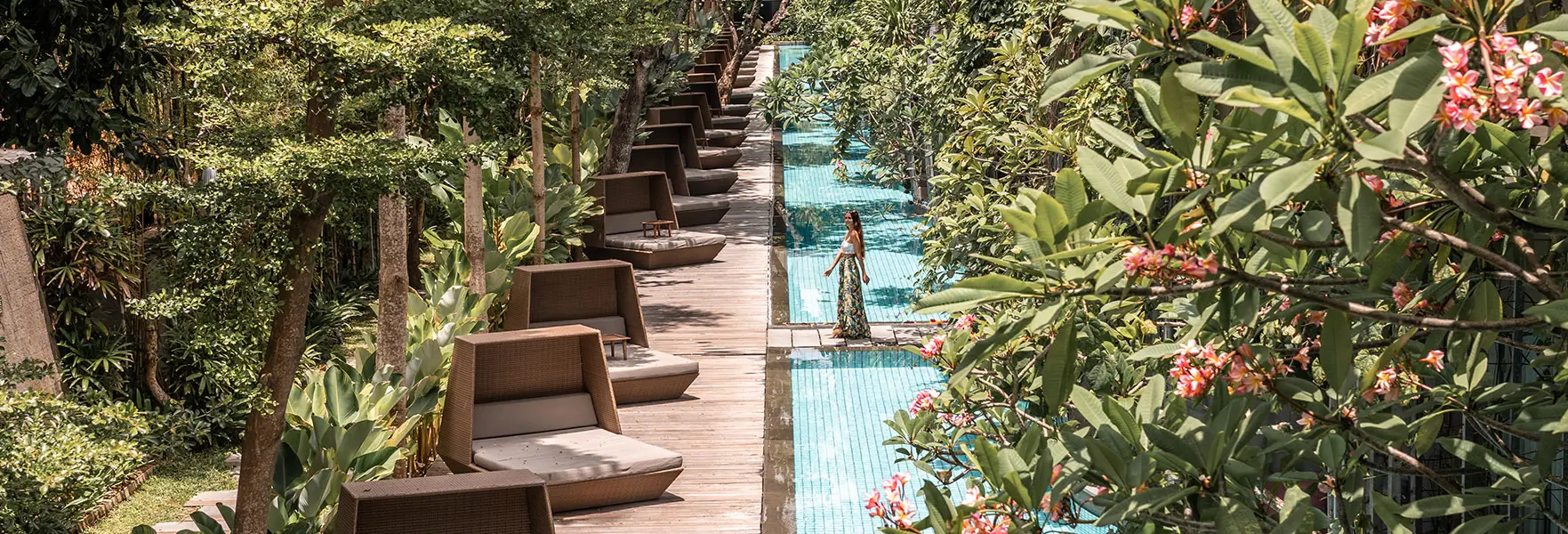 Maya Resort Sanur pathway, lined with modern lounge chairs and surrounded by lush greenery and vibrant flowers, adjacent to a narrow, clear swimming pool. A woman in a floral skirt walks by the pool, enjoying the serene environment.