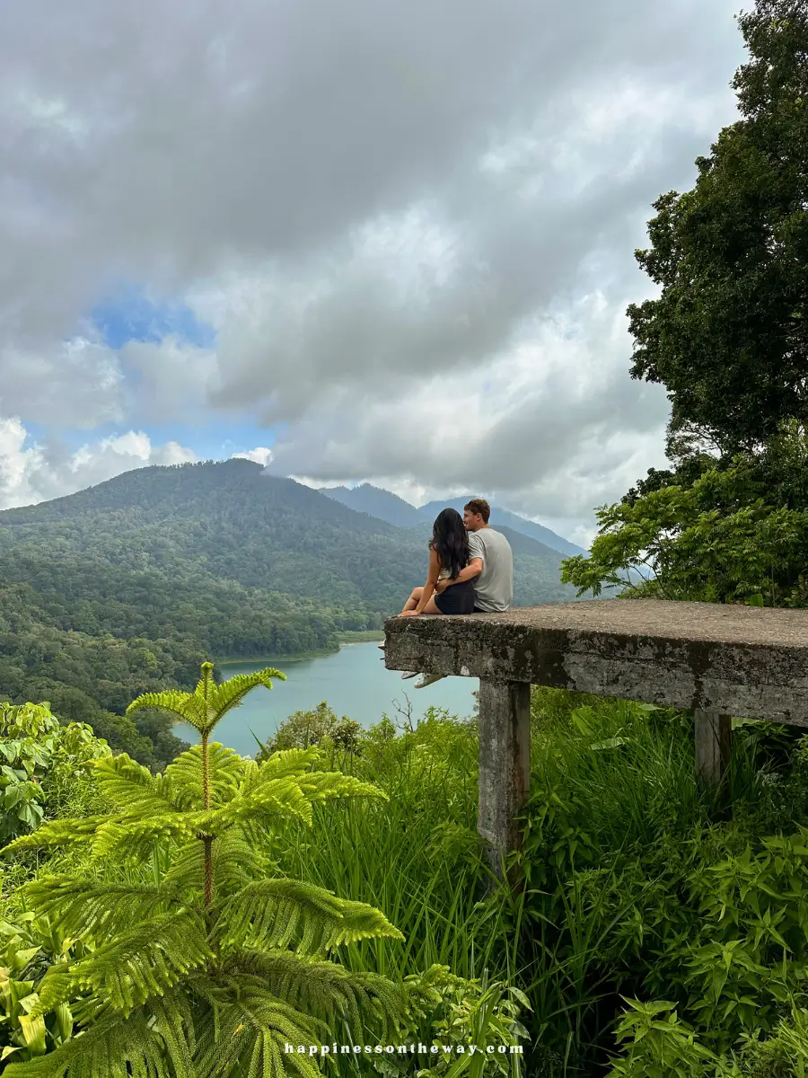 Me and my partner sitting on the edge of a concrete platform overlooking a lush green valley with a lake and mountains in Munduk, Bali. The sky is overcast, adding a serene and introspective mood to the scene.