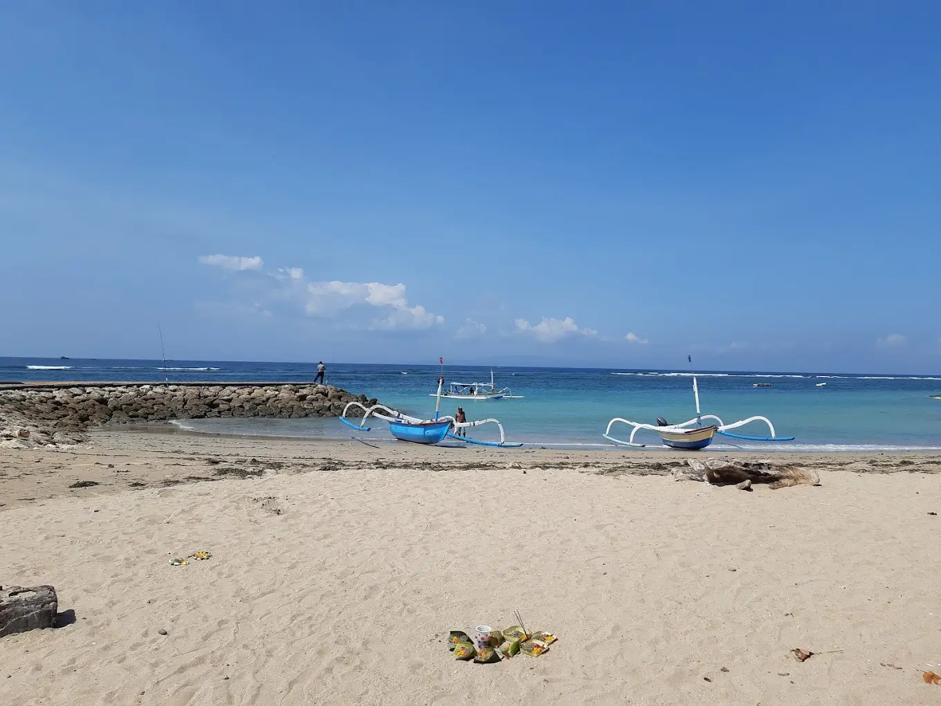 A serene view of Pantai Bangsal Sanur with traditional blue fishing boats resting on the sandy beach, a rocky jetty extending into the clear turquoise water, and a fisherman standing at the end under a bright blue sky.