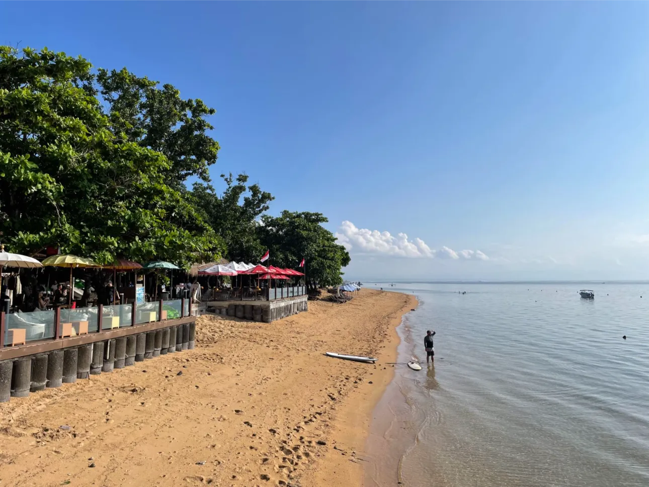 Restaurants with sun umbrellas in front of Pantai Duyung Beach with a person on the shore and a paddleboard.