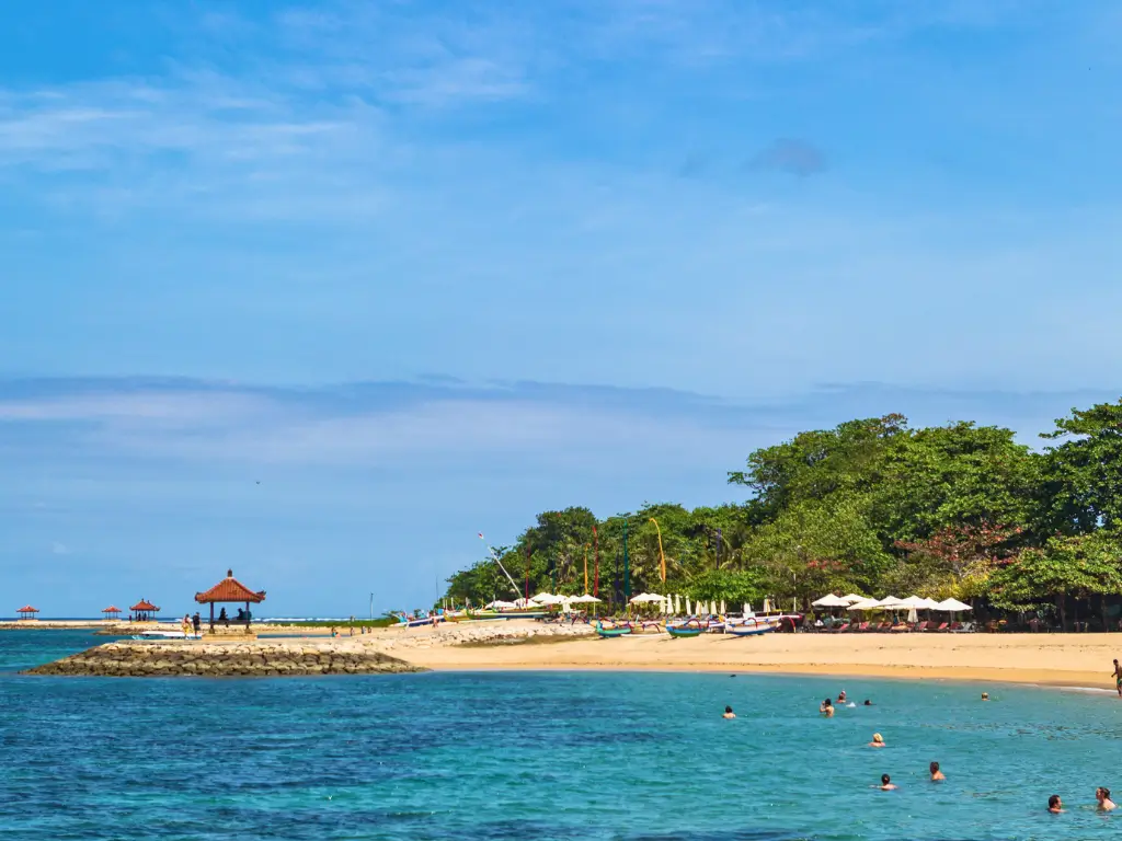 Sanur Beach in Bali with clear turquoise water, a sandy shore, and people swimming. A small pavilion stands on a rocky jetty, and green trees line the beach in the background.