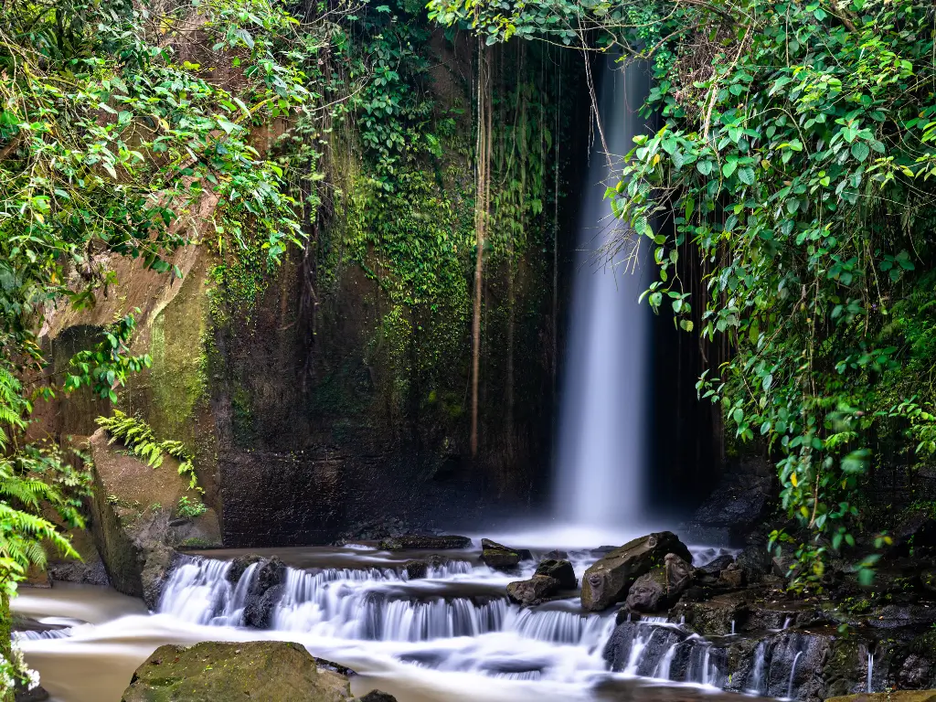 Sumampan Waterfall cascading over a rocky ledge into a calm, forested pool surrounded by green vegetation. 