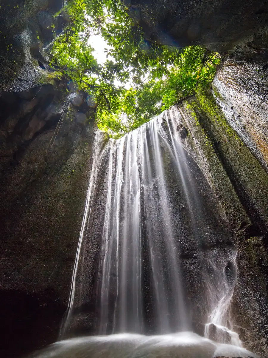 Tukad Cepung Waterfall flows gently down a rocky crevice, with sunlight streaming through the lush green foliage above. The water descends into a secluded pool.