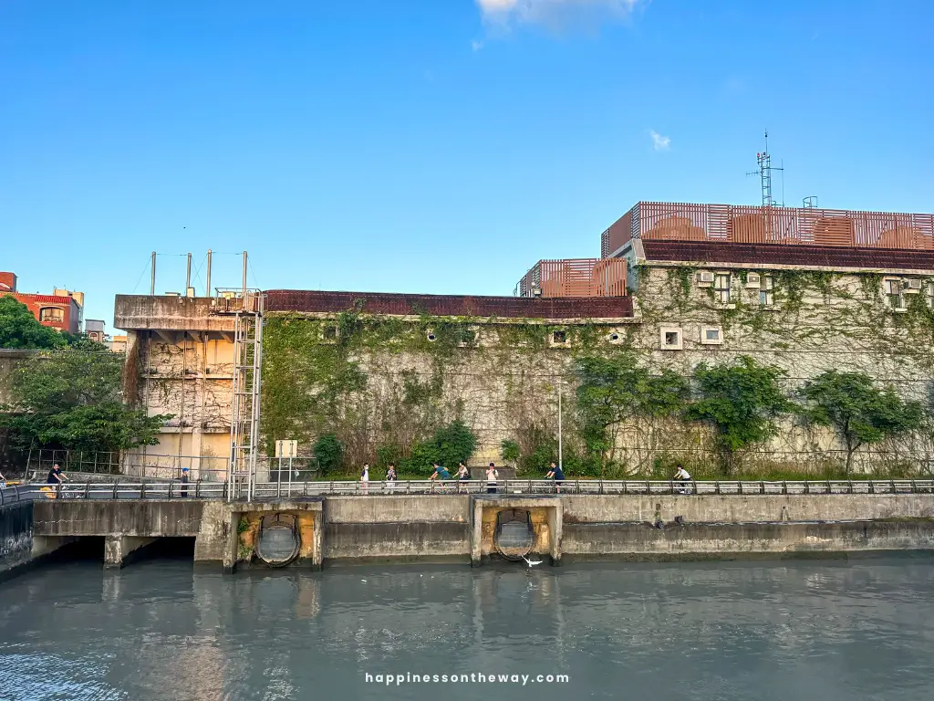 An old concrete building covered with green ivy and moss along the riverside at Dadaocheng Pier Plaza. A bicycle path runs in front of the building, with several people walking and cycling.