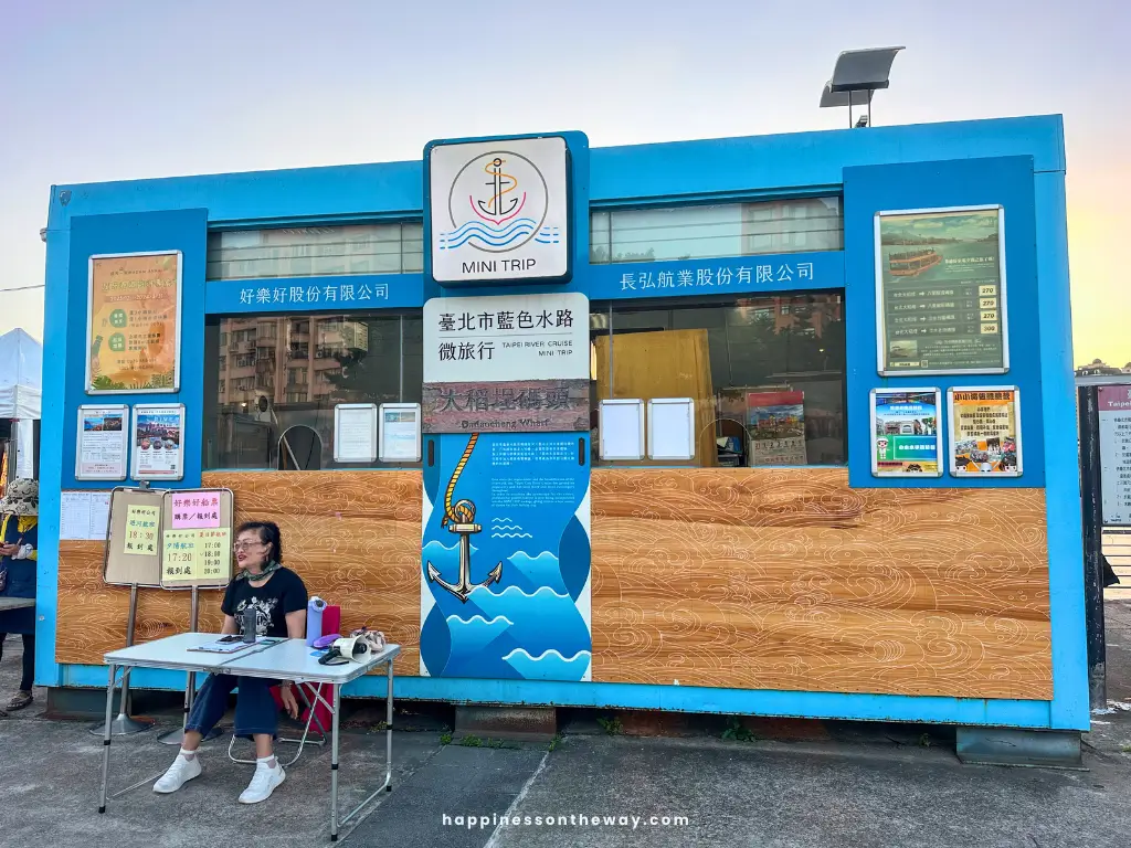 A booth for 'Mini Trip' boat tours at Dadaocheng Pier Plaza, featuring various posters and information about Taipei river cruises. A woman is seated at a table in front of the booth.