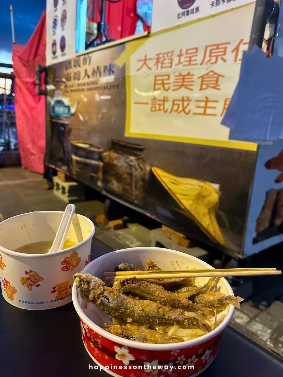  A bowl of deep-fried fish sticks served with wooden skewers next to a cup of soup at a food stall in Dadaocheng Pier Plaza night market. The background shows a brightly lit stall with signs in Chinese characters.