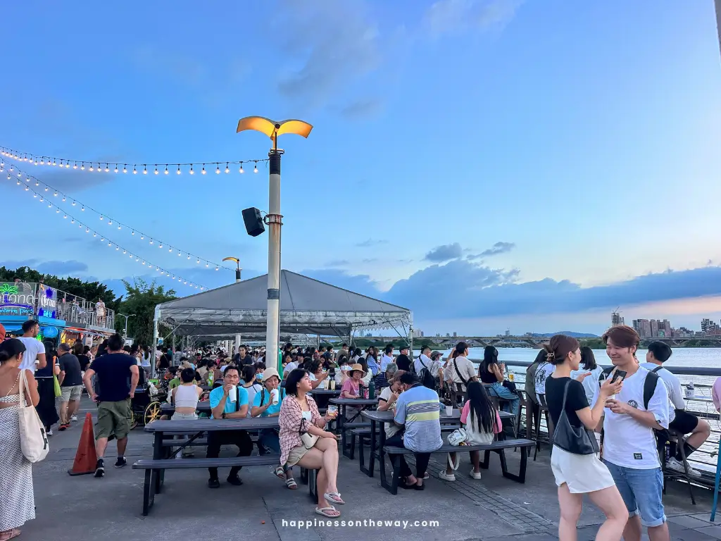 Crowds of people seated and walking around Dadaocheng Pier Plaza, enjoying the night market by the riverside. The sky is clear with string lights hanging above, creating a festive atmosphere.