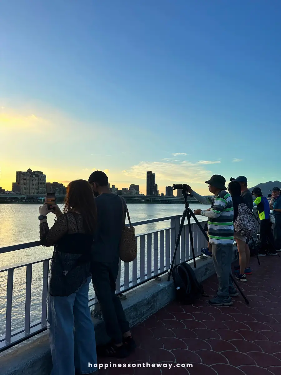 People with cameras and smartphones take photos of the sunset over the river at Dadaocheng Pier Plaza. The sky is clear with a gradient from golden to blue, and the city skyline is visible in the background.