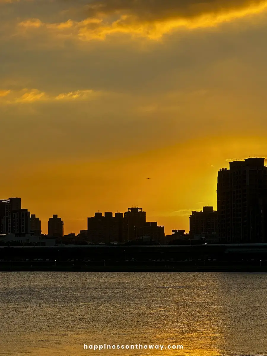 Sunset over the Taipei skyline with the water in the foreground from Dadaocheng Pier Plaza, featuring a warm orange and yellow sky with clouds.