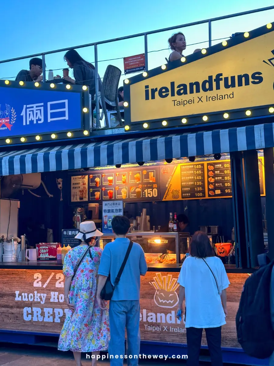 People standing in front of the 'irelandfuns Taipei X Ireland' food stall at Dadaocheng Pier Plaza night market. The stall has a menu with various food items displayed, and a seating area on the upper level.