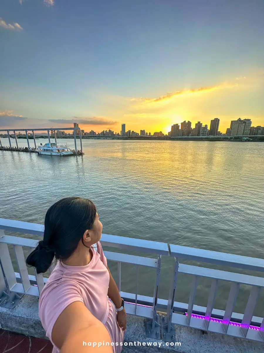 Me in a pink shirt takes a selfie at Dadaocheng Pier, with a scenic view of the river and city skyline during sunset. A boat is docked at the pier, and the sky is lit with golden hues as the sun sets behind the buildings.