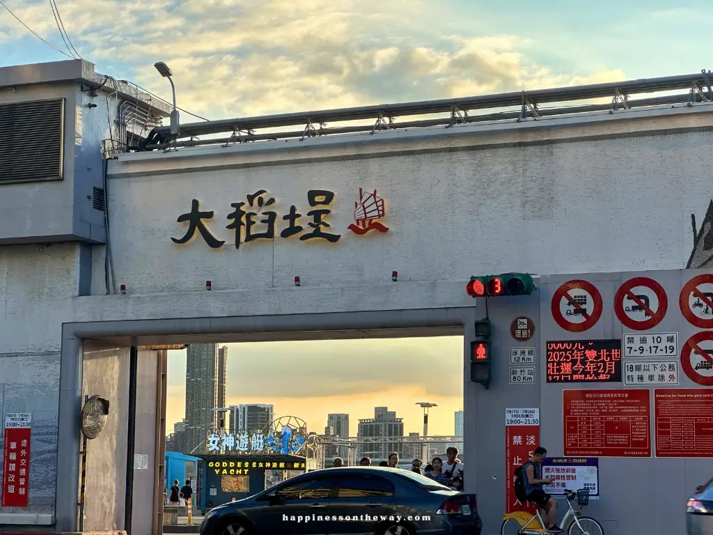 An entrance gate of Dadaocheng Wharf with large Chinese characters on a white wall, "大稻埕" (Dadaocheng), and smaller signage including "Goddess Yacht" visible through the gate.