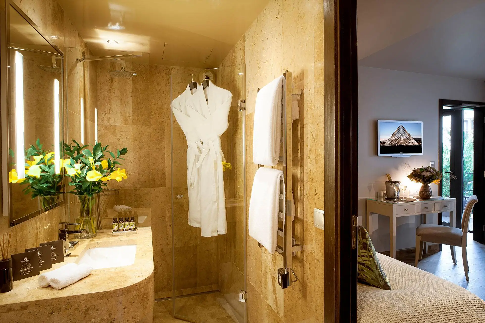 Bathroom at B Montmartre Hotel featuring beige marble walls, a walk-in shower with two white bathrobes, and a heated towel rack with fresh towels.