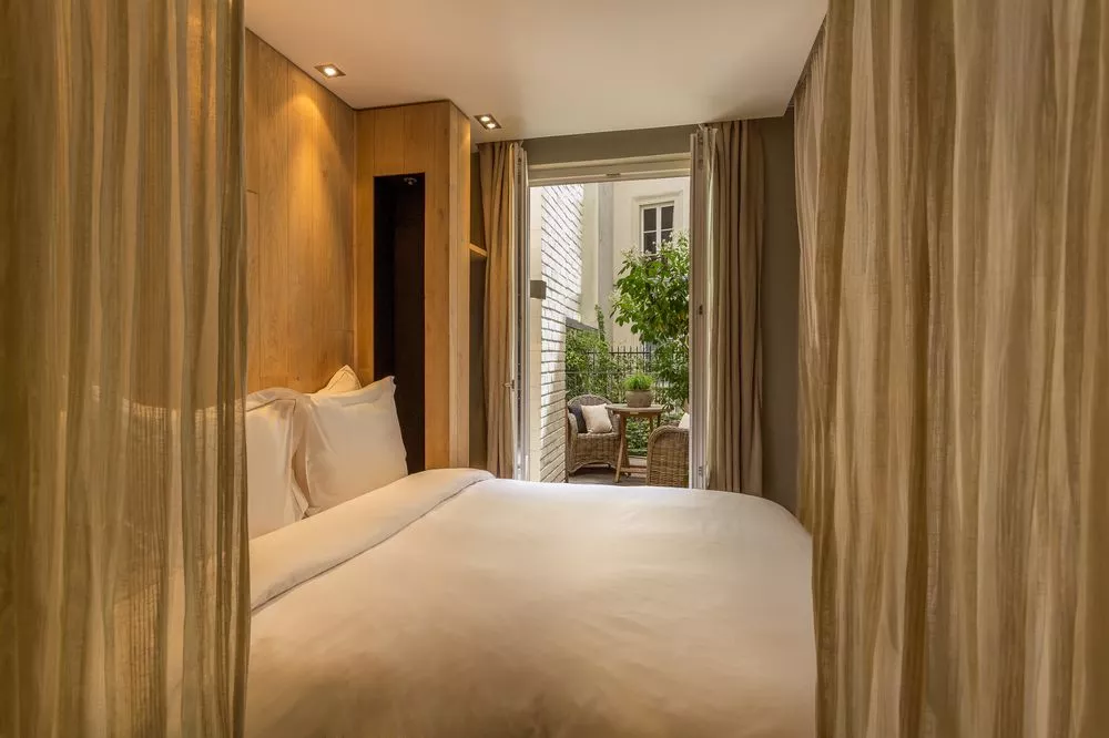 Cozy room at the Hidden Hotel in Paris featuring white linens, brown curtains, and an inviting garden balcony with chairs and tables.