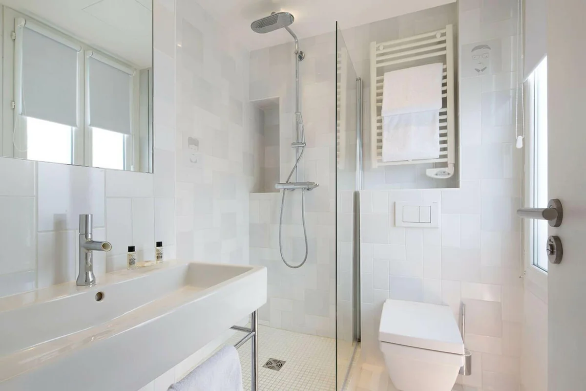 A white-tiled bathroom with a sleek modern design at the quiet Hotel Mistral in Paris, featuring a large mirror, clean lines, and simple elegance.