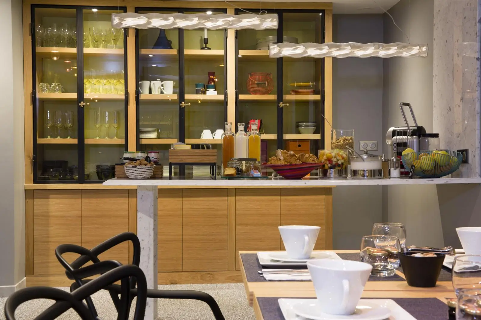 The breakfast area of Hôtel Signature Saint Germain des Prés, a quiet hotel in Paris, with neatly set tables and a buffet rack in the background displaying fresh glassware, croissants, fruit, yogurt, and toast