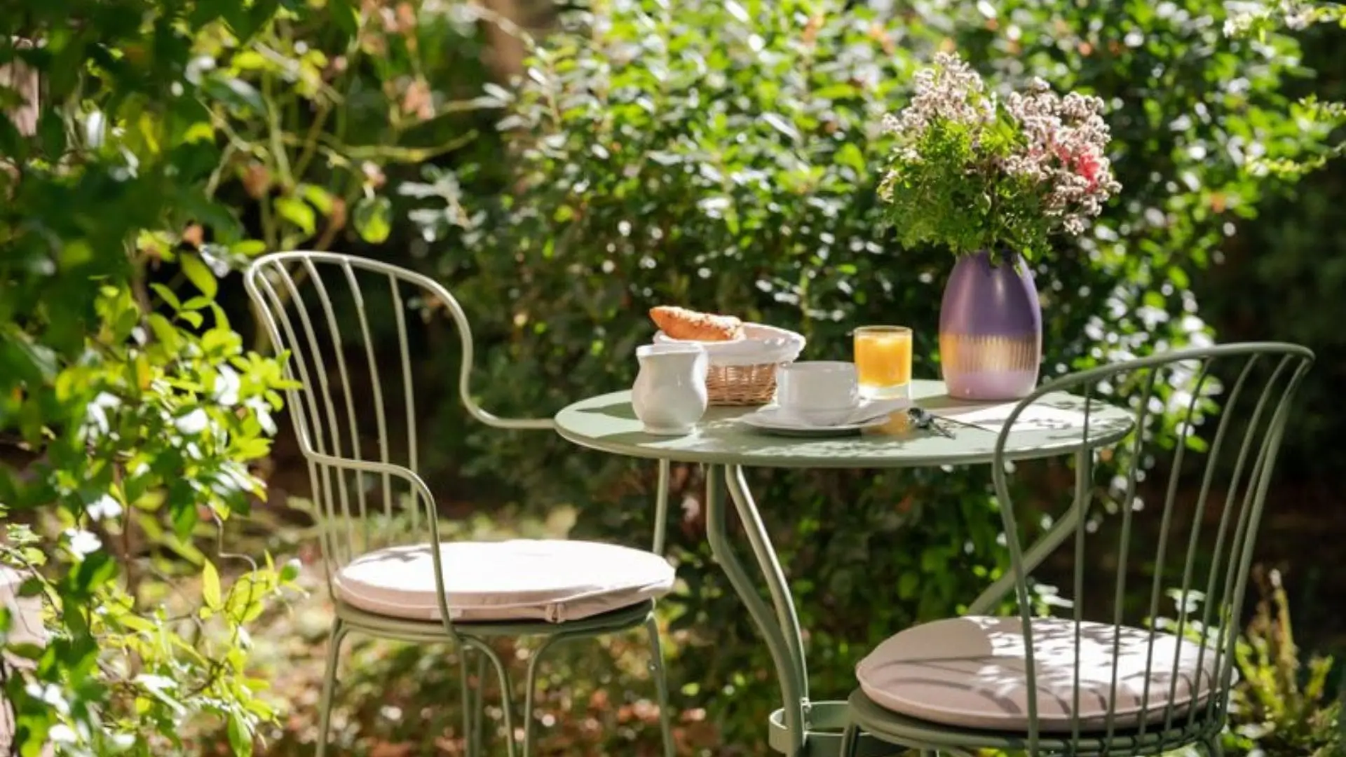 A peaceful breakfast setting at Hotel des Grandes Ecoles, featuring a set table with two chairs, fresh orange juice, coffee with milk, and a basket of bread nestled in a tranquil garden.