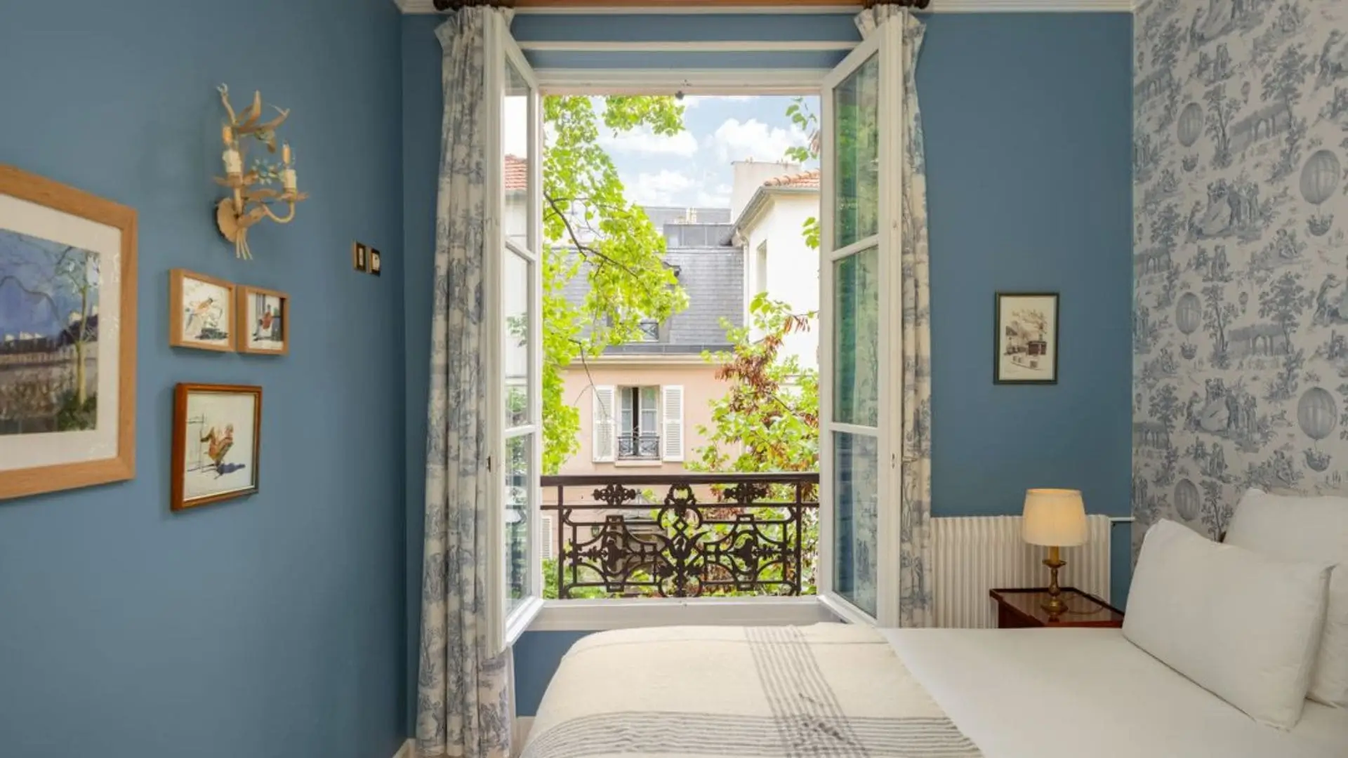 The serene bedroom at Hôtel Le Relais Montmartre showcases a white and blue interior with a large window overlooking a peaceful garden