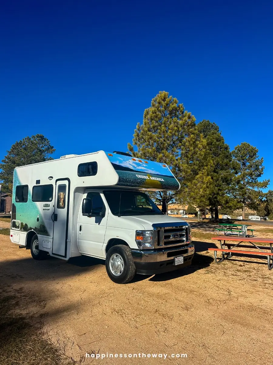 A white Cruise America RV parked in Ruby's Inn RV Park under a clear blue sky, with tall trees in the background.