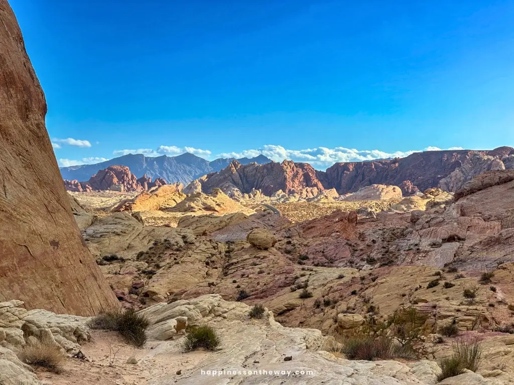 A panoramic view from Rainbow Vista in Valley of Fire State Park, featuring layered sandstone formations in shades of red, orange, and cream, set against a backdrop of distant mountains under a clear blue sky.