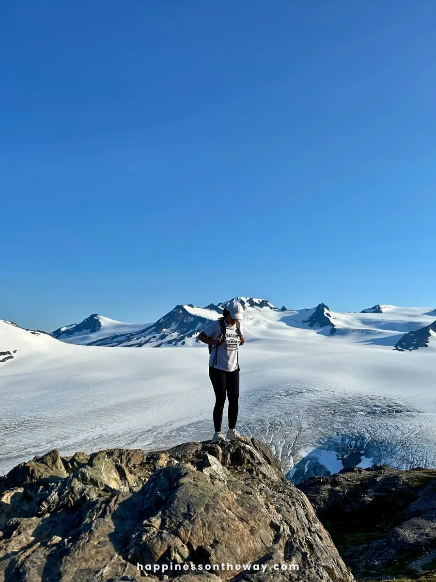 I am wearing a white cap, t-shirt, and black leggings stands on a rocky peak overlooking a vast glacier and snow-capped mountains under a clear blue sky. 