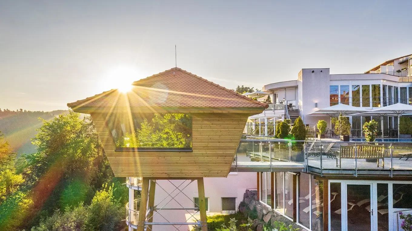 Scenic view of Hotel Kronelamm featuring a modern glass-walled building with outdoor terraces and a unique elevated wooden treehouse suite.